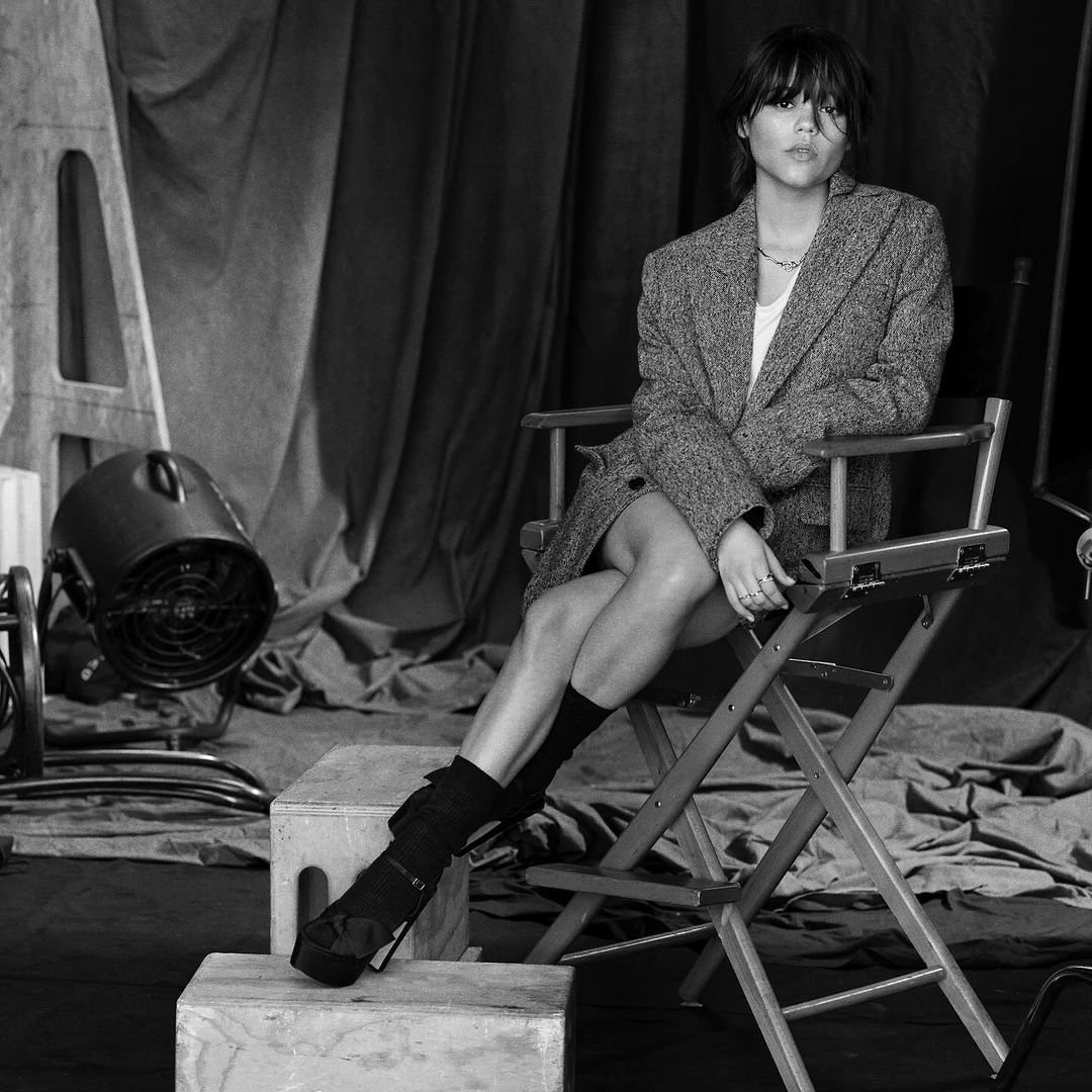 Black and white portrait of a young woman with short hair wearing a tweed blazer, seated in a director's chair in a photo studio.