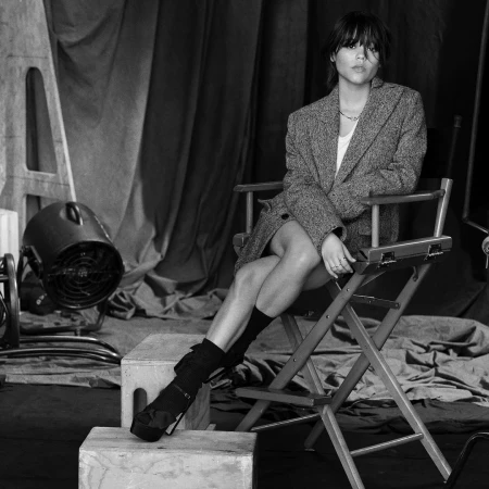 Black and white portrait of a young woman with short hair wearing a tweed blazer, seated in a director's chair in a photo studio.