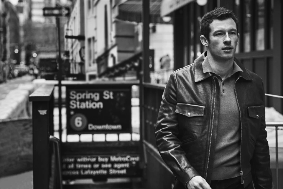 Black and white medium shot of a man in a leather jacket standing outside of the Spring Street subway station in New York City.