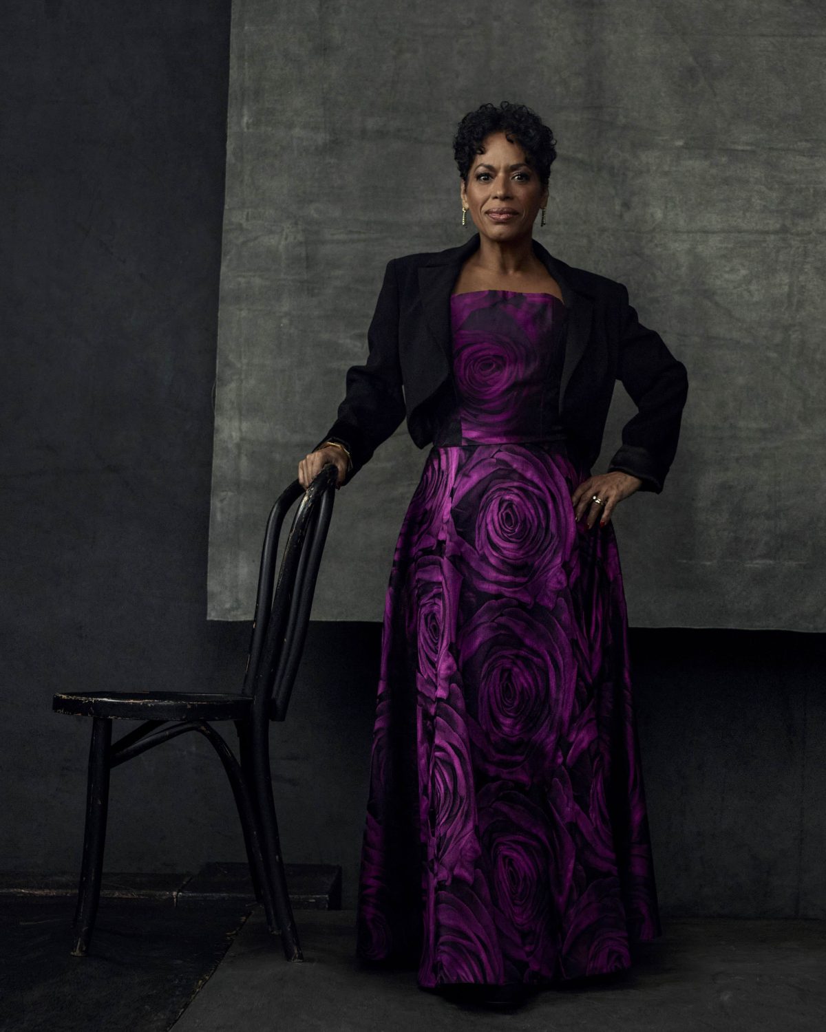 Elegant adult woman in floral gown and black jacket standing indoors with dark gray background, posing and holding a black chair.