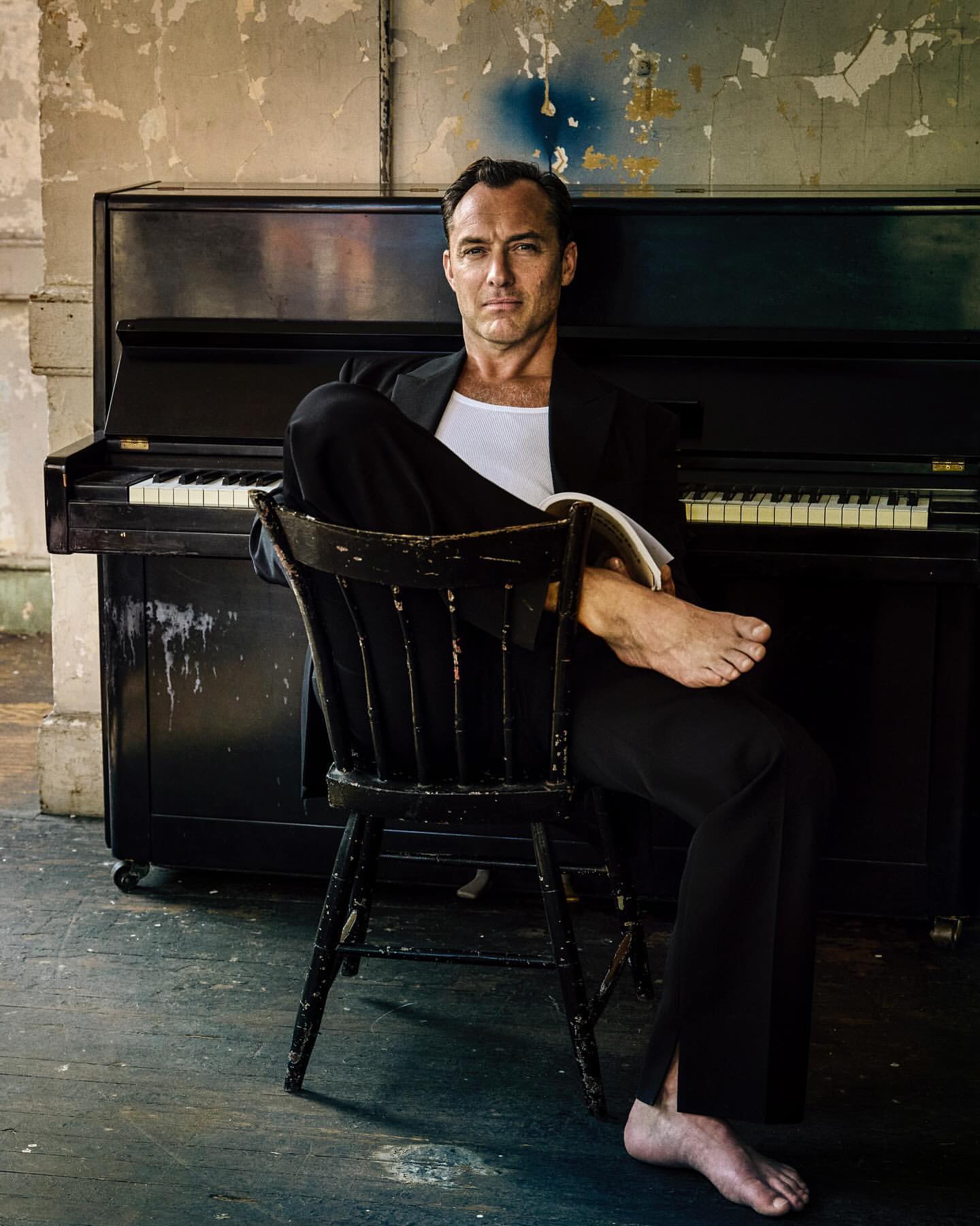 Portrait of Jude Law seated barefoot on a wooden chair in front of an upright piano wearing a black suit and white tank top.