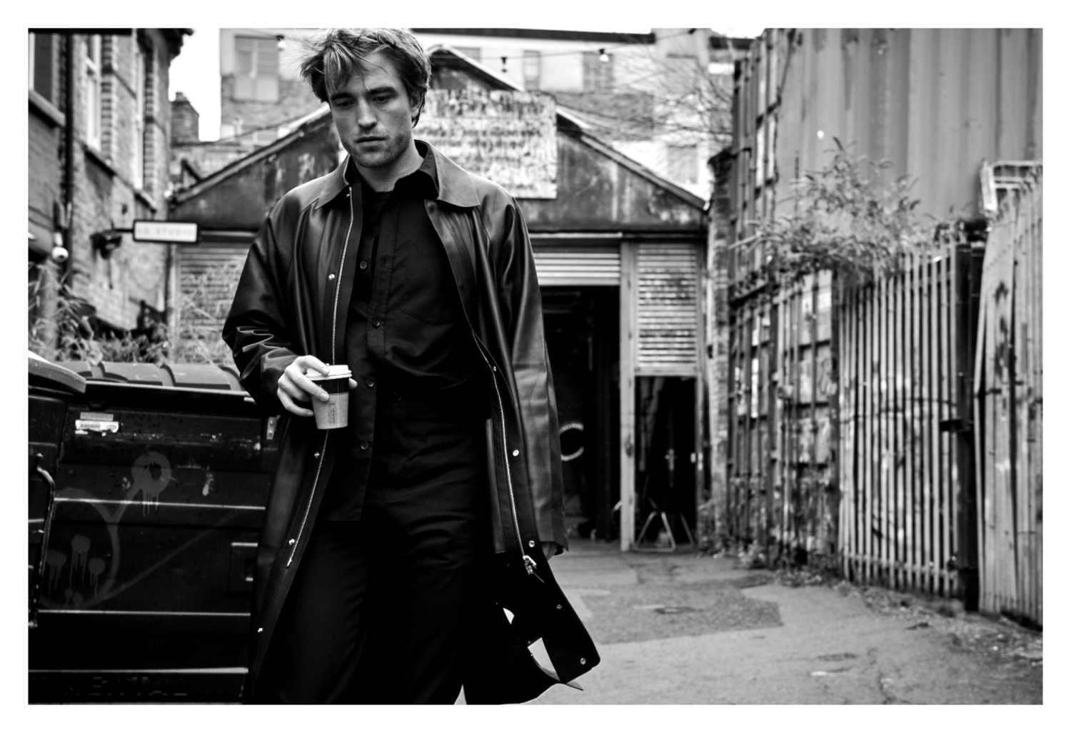 Black-and-white outdoor shot of a man in a coat holding a coffee cup, walking down a city alleyway with metal buildings.