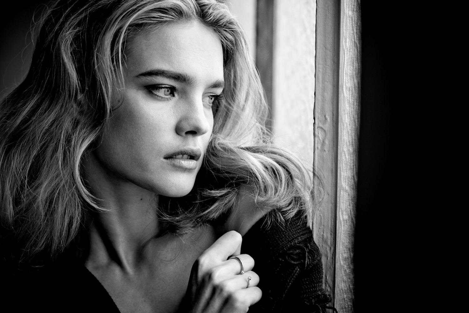Close-up black and white portrait of a woman looking out a window wearing dark knitwear with rings on her fingers.