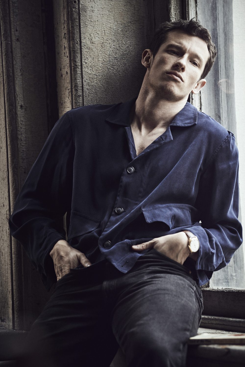 A man wearing a blue button-up shirt and dark trousers leaning against a textured interior wall by a window.