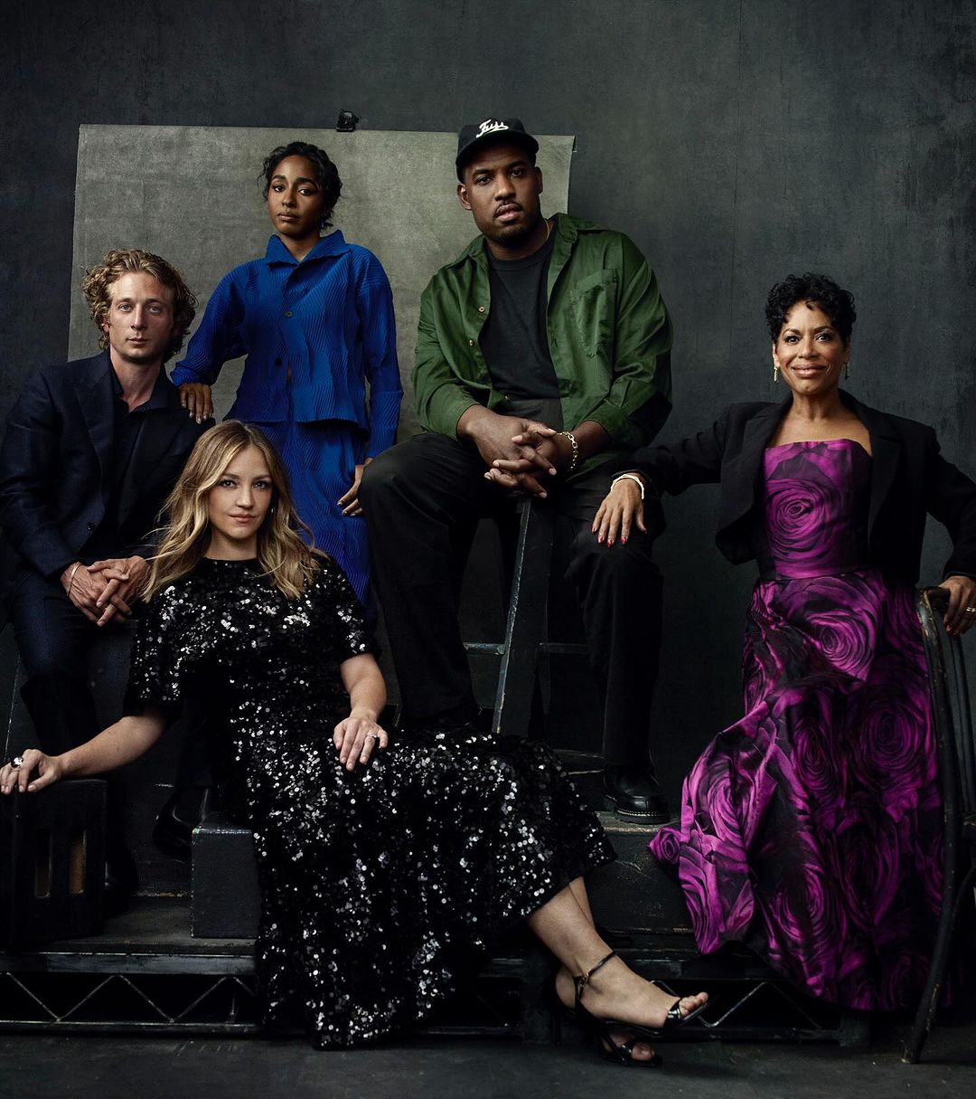Group portrait of five diverse adults in a photo studio, wearing formal and casual clothing, posing against a gray backdrop with soft lighting.