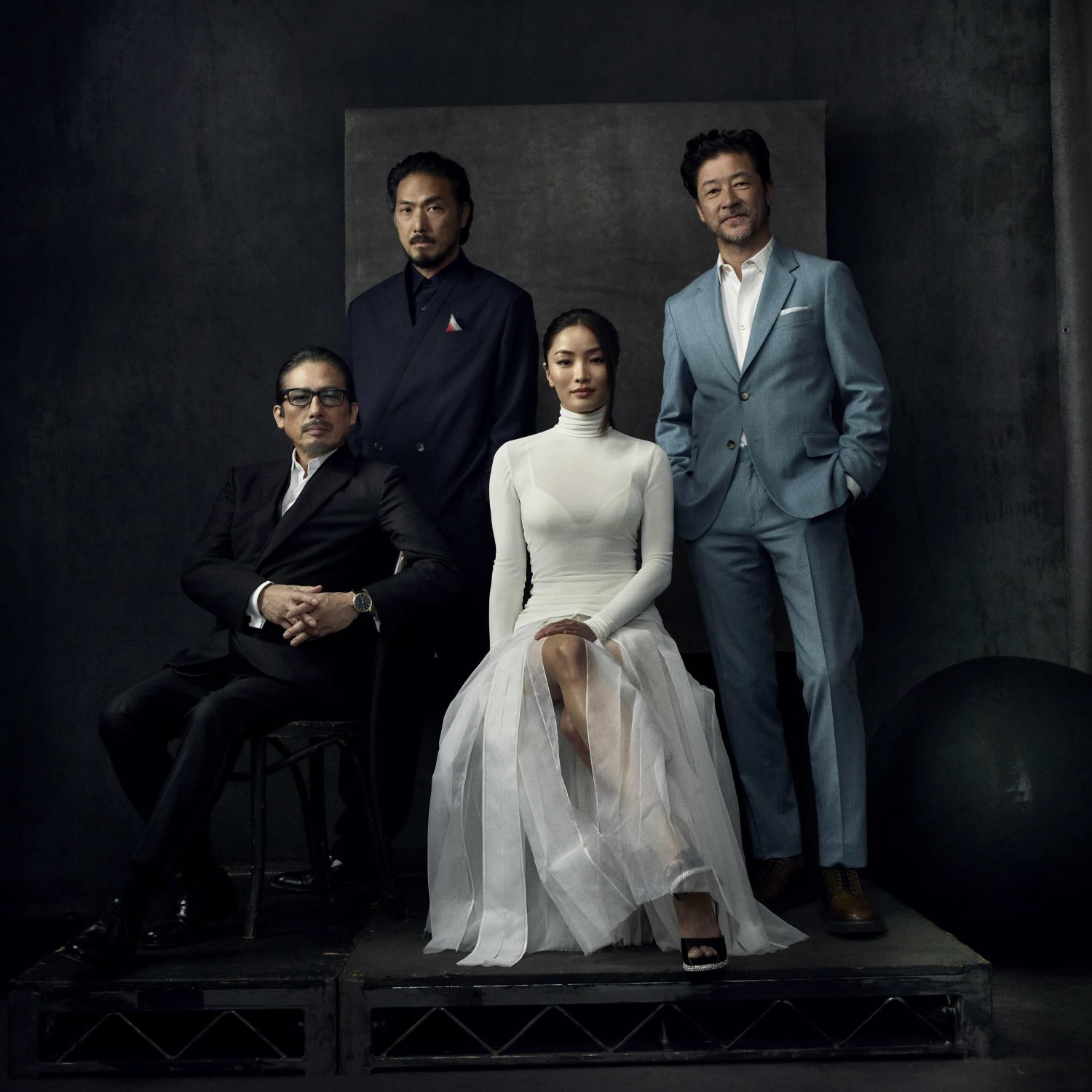 Group portrait of four Asian adults, three men and one woman, formally dressed in suits and a white dress in a studio setting.