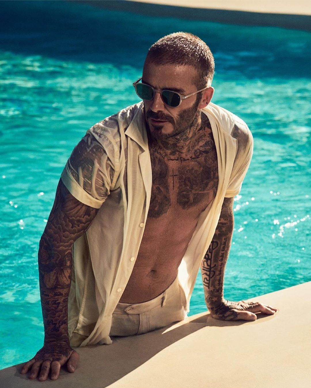 David Beckham in a swimming pool wearing sunglasses and an unbuttoned shirt, showing tattoos. Outdoor, daytime setting.