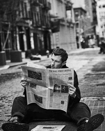 Man in black jacket and jeans sits on street reading Wall Street Journal newspaper in black and white with buildings in background.
