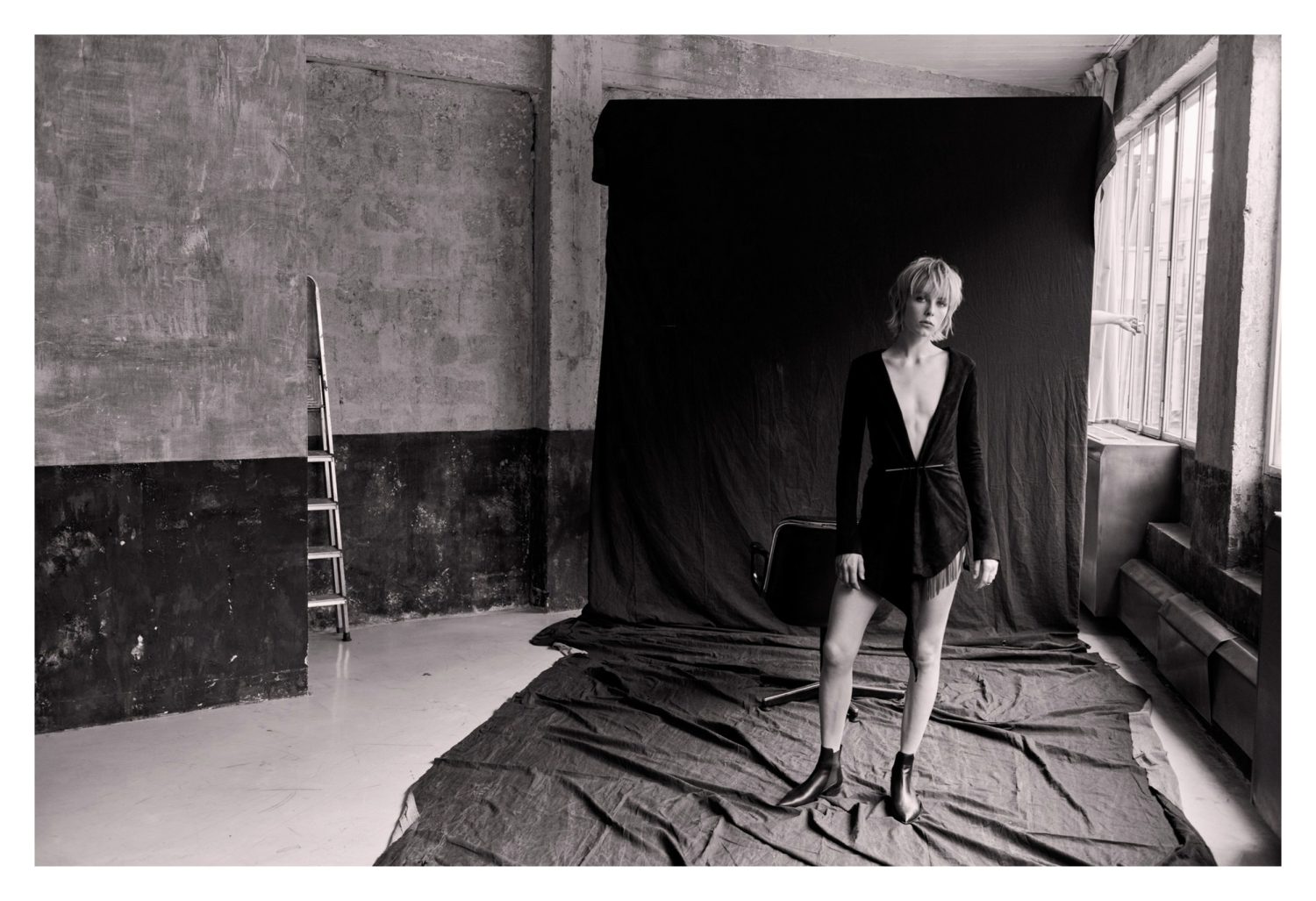 Model in dark wrap dress and boots standing before a black backdrop in a warehouse studio by Matthew Brookes