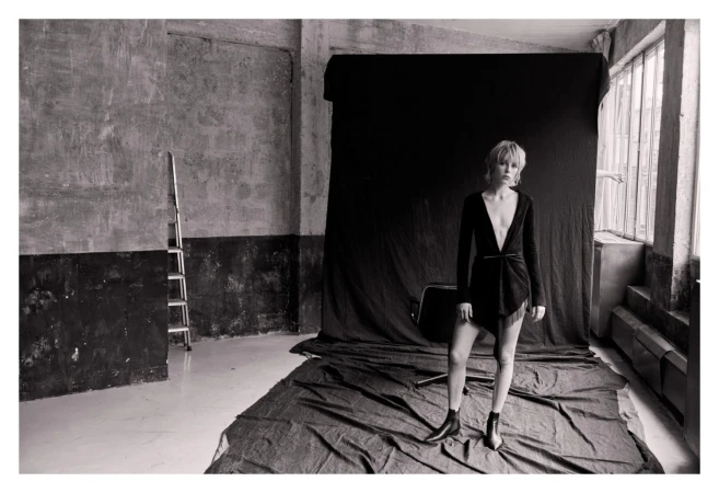 Model in dark wrap dress and boots standing before a black backdrop in a warehouse studio by Matthew Brookes