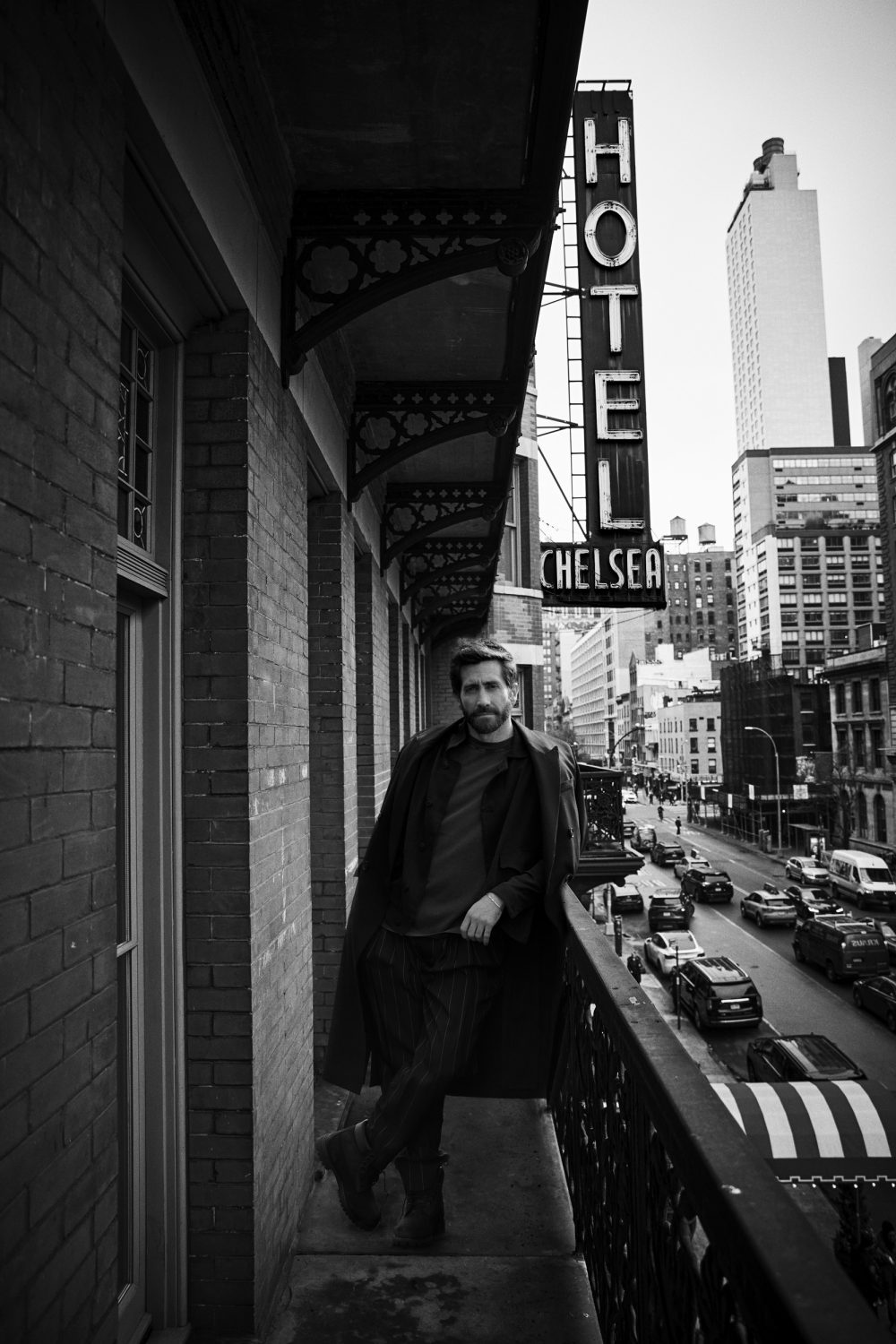 Black and white portrait of a man standing on a balcony with an urban street scene in the background and HOTEL CHELSEA sign visible.