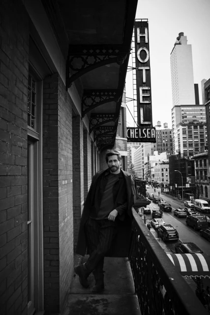 Black and white portrait of a man standing on a balcony with an urban street scene in the background and HOTEL CHELSEA sign visible.