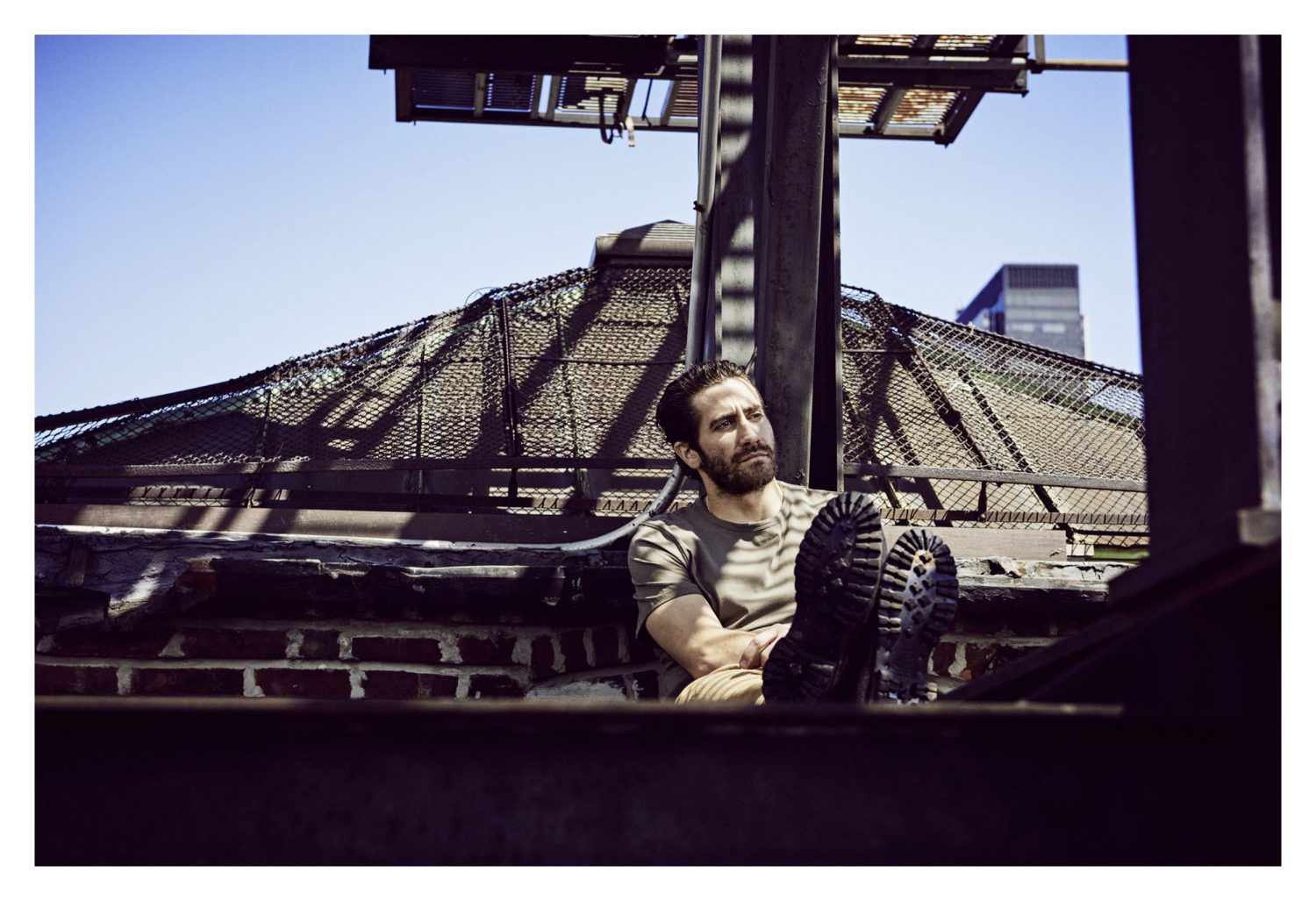 Bearded adult male in casual clothing lounges on a rooftop with city buildings against a sunny blue sky, wearing work boots.