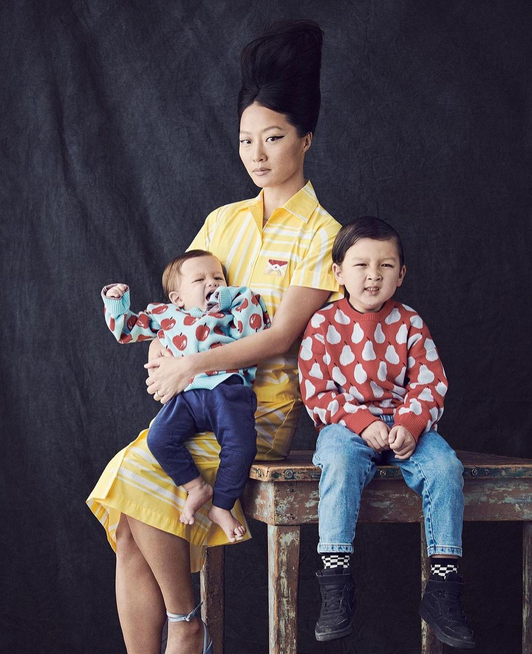Asian woman with a high hairstyle poses indoors with two young children, one infant and one toddler, wearing patterned sweaters and jeans.