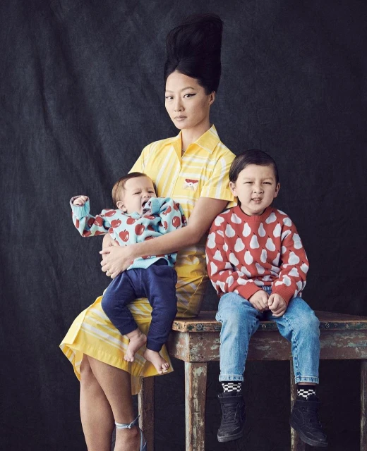 Asian woman with a high hairstyle poses indoors with two young children, one infant and one toddler, wearing patterned sweaters and jeans.