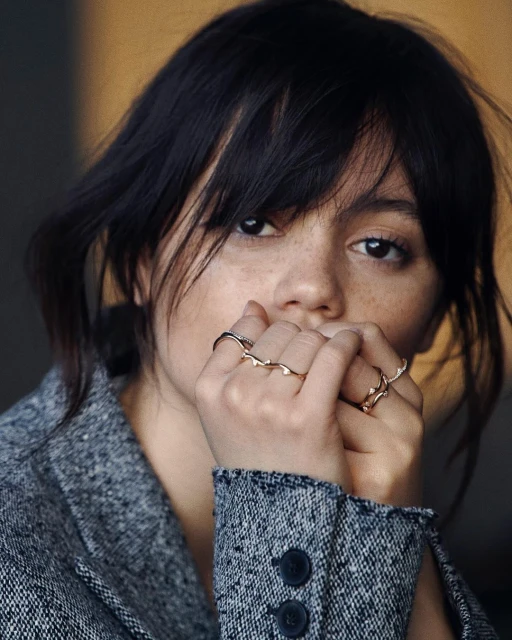 Close-up portrait of a young adult female with dark hair and freckles, wearing rings and a grey jacket, against a blurred background.
