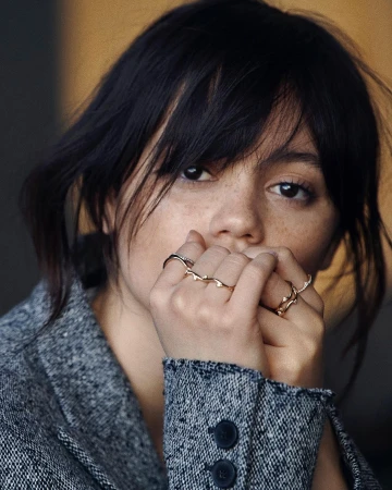 Close-up portrait of a young adult female with dark hair and freckles, wearing rings and a grey jacket, against a blurred background.