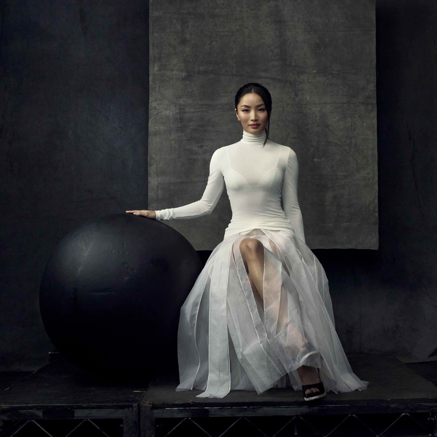 Elegant woman in white turtleneck and sheer skirt poses with large black ball in a dark photo studio setting with artistic backdrop.