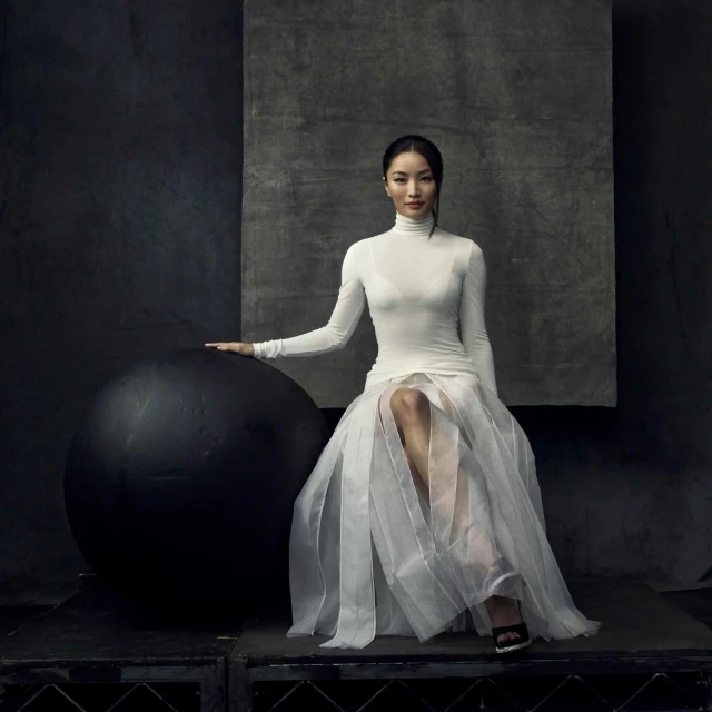 Elegant woman in white turtleneck and sheer skirt poses with large black ball in a dark photo studio setting with artistic backdrop.