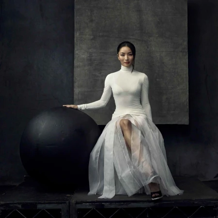 Elegant woman in white turtleneck and sheer skirt poses with large black ball in a dark photo studio setting with artistic backdrop.