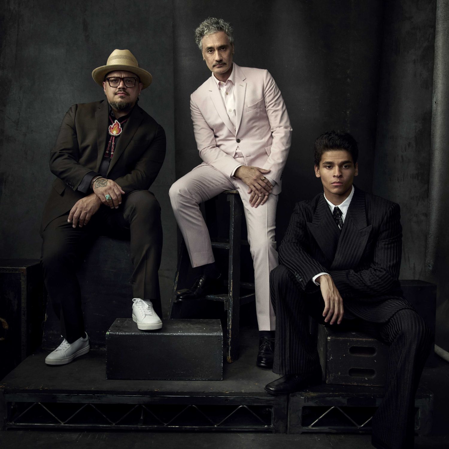 Portrait of three stylishly dressed men, one seated on a black box, against a dark grey studio backdrop, medium shot, fashion editorial.