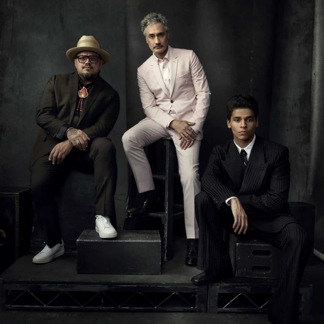 Portrait of three stylishly dressed men, one seated on a black box, against a dark grey studio backdrop, medium shot, fashion editorial.