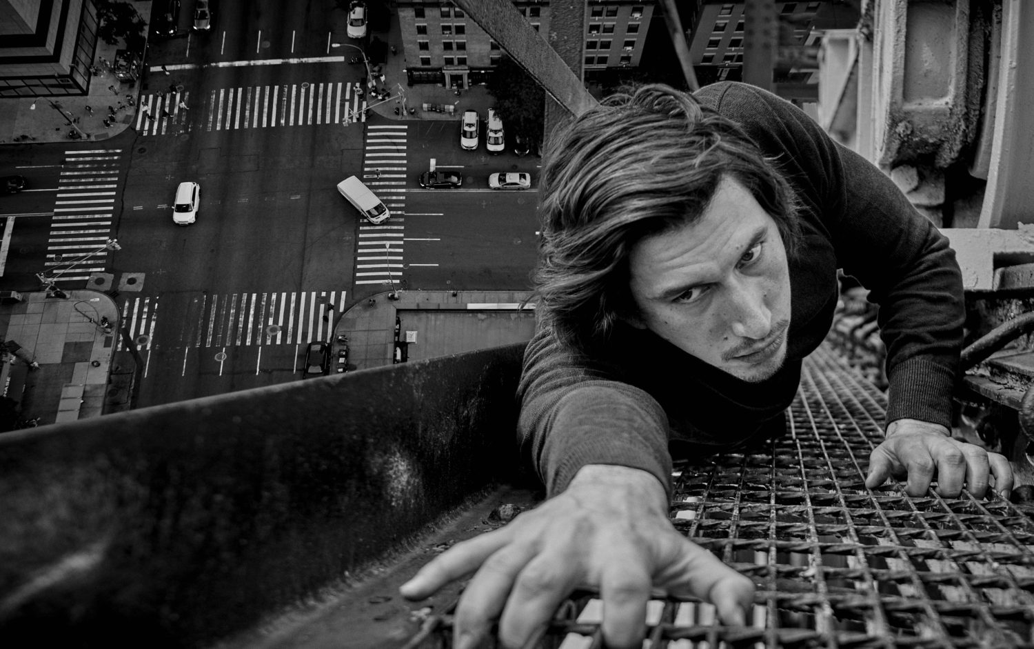 Black and white, low-angle image of an adult male clinging to a rooftop, with an urban street scene visible below, shot from above.