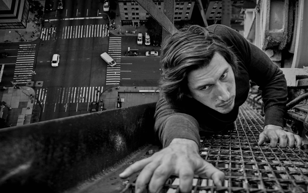 Black and white, low-angle image of an adult male clinging to a rooftop, with an urban street scene visible below, shot from above.