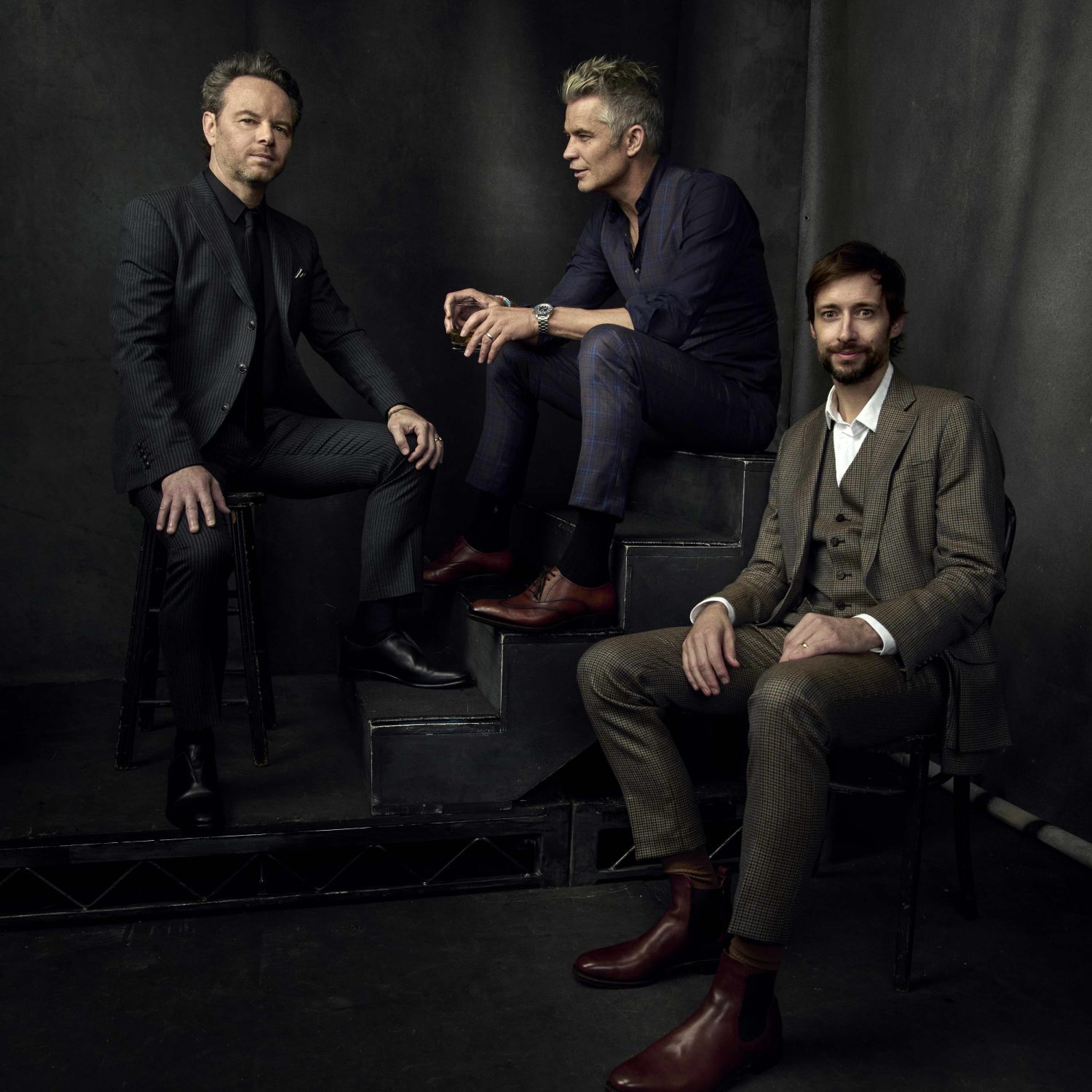 Three men in stylish suits pose indoors. One man sits on steps with the other two positioned around him in a studio setting.