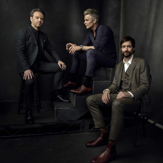 Three men in stylish suits pose indoors. One man sits on steps with the other two positioned around him in a studio setting.