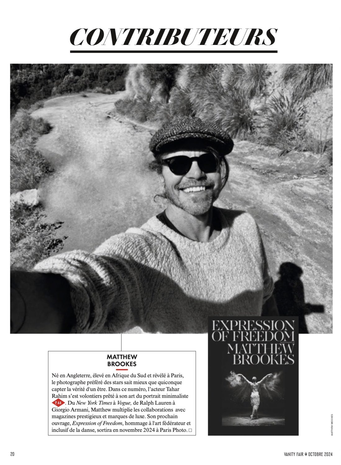 Black and white outdoor portrait of Matthew Brookes wearing sunglasses and a cap, smiling in front of a pale textured background.