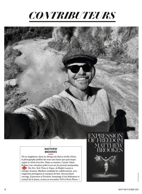 Black and white outdoor portrait of Matthew Brookes wearing sunglasses and a cap, smiling in front of a pale textured background.