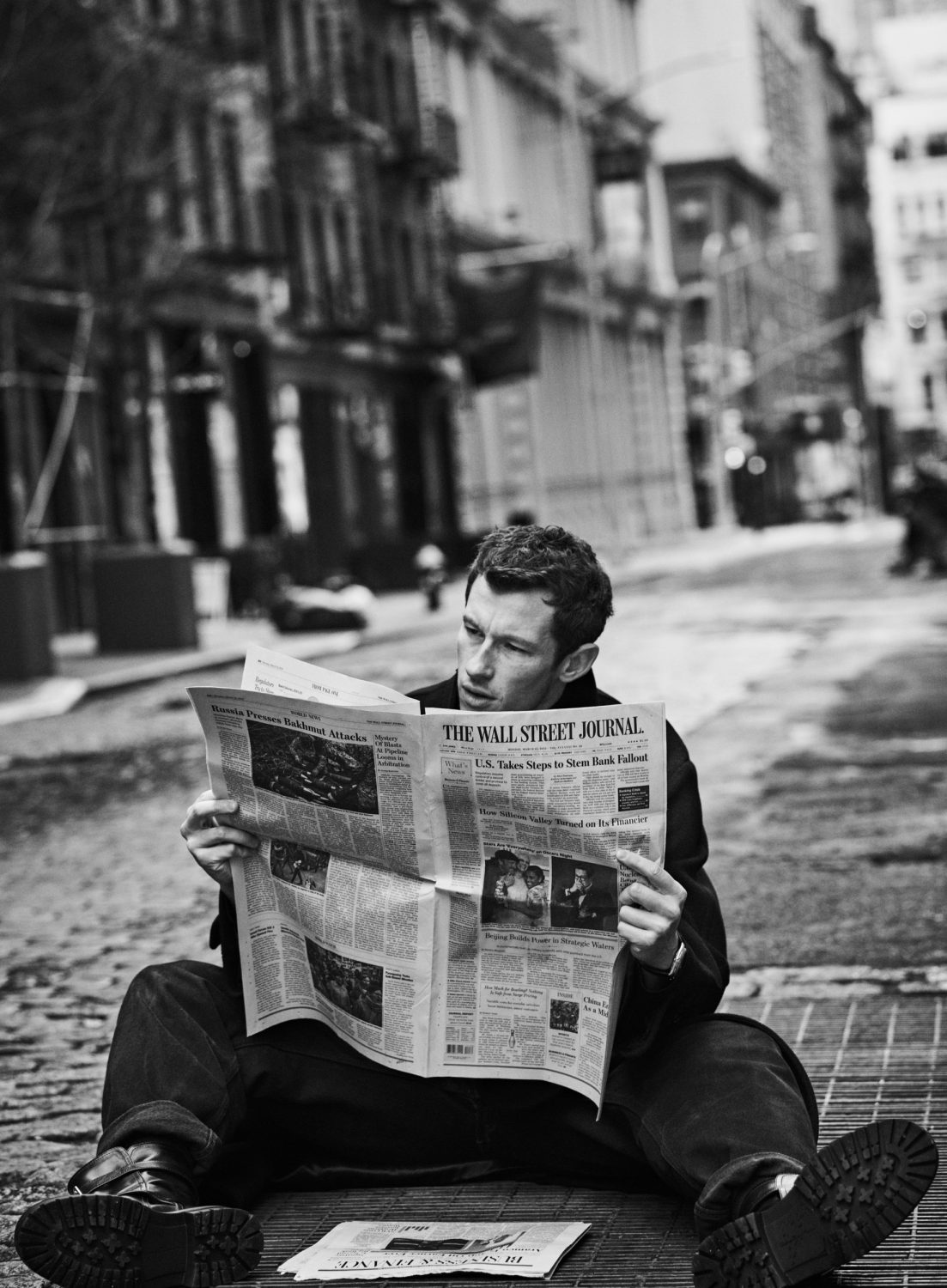 Monochrome image of a man sitting on a cobblestone street reading The Wall Street Journal newspaper, with buildings in the background.