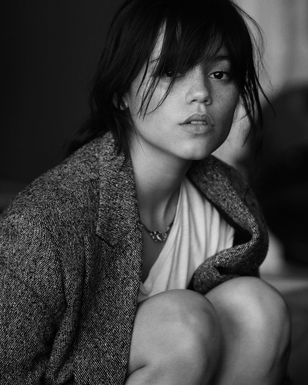 Black and white portrait of a young adult woman with dark hair wearing a blazer and necklace. She is crouching and looking up.