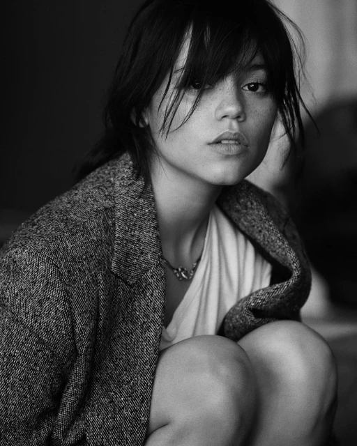 Black and white portrait of a young adult woman with dark hair wearing a blazer and necklace. She is crouching and looking up.