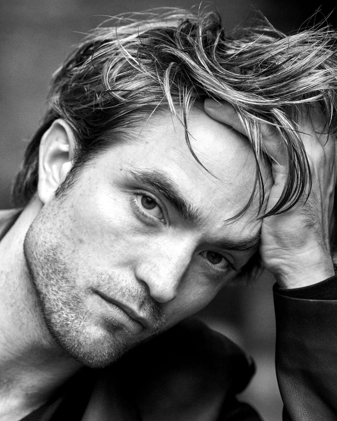 Black and white portrait of a man with light hair, head resting on his hand, looking at the camera. Moody studio lighting. 