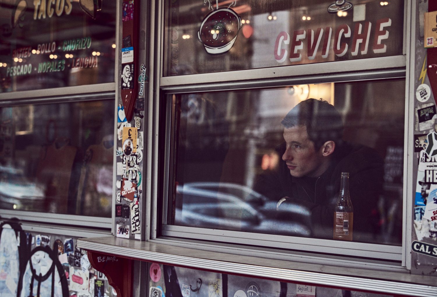 Adult male looks out window of a taco shop; the eatery advertises ceviche. Stickers adorn the window frame. The photo captures an urban scene.