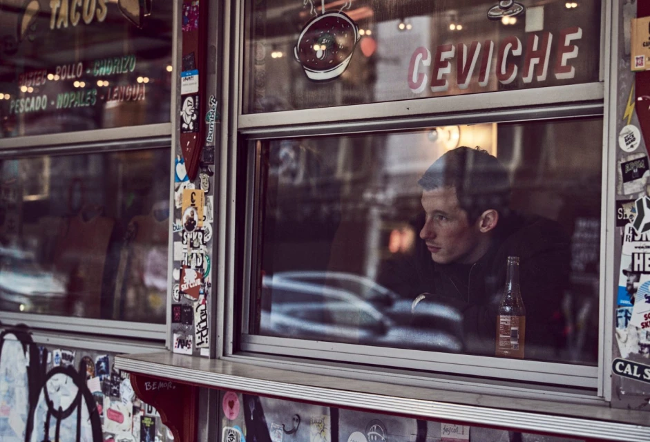 Adult male looks out window of a taco shop; the eatery advertises ceviche. Stickers adorn the window frame. The photo captures an urban scene.