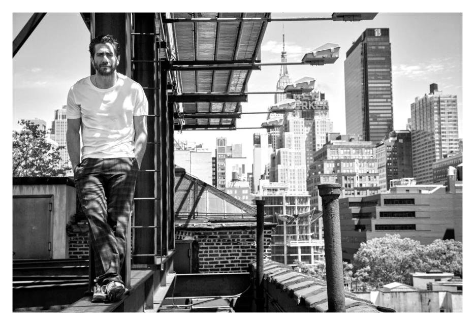 Black and white photo of an adult male in a white t-shirt and patterned pants standing on a rooftop with a cityscape background.