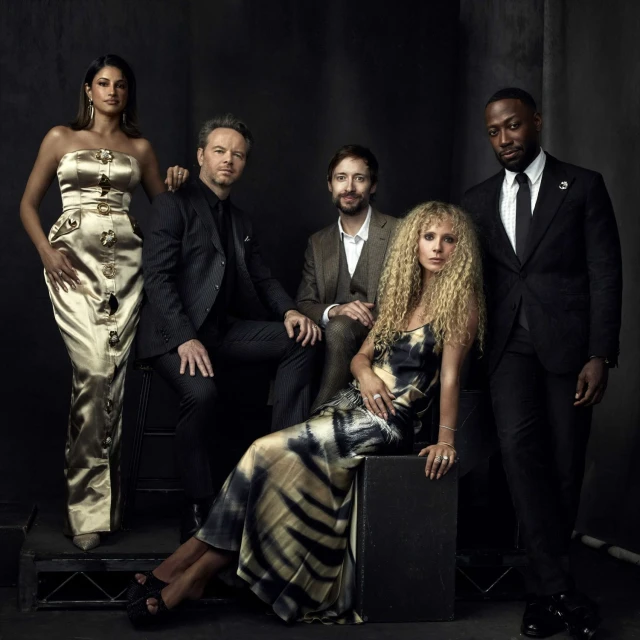 Group portrait of five actors in formal attire posing in front of a dark backdrop in a studio setting with warm, soft lighting.
