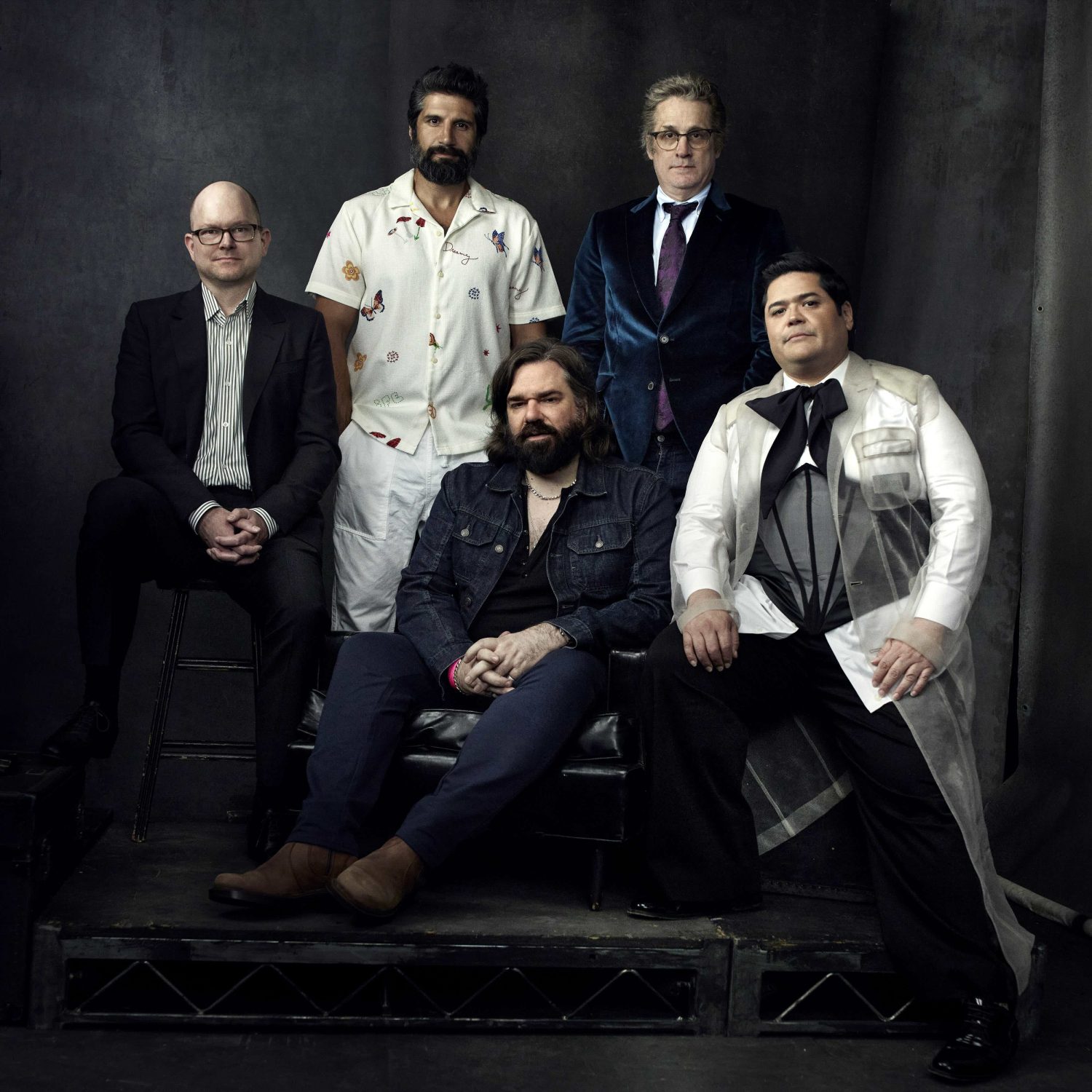 Group portrait of five adult males seated on a stage with a dark backdrop, wearing suits and casual wear in an indoor setting.