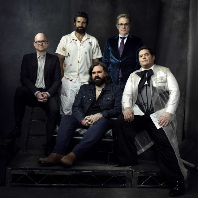 Group portrait of five adult males seated on a stage with a dark backdrop, wearing suits and casual wear in an indoor setting.