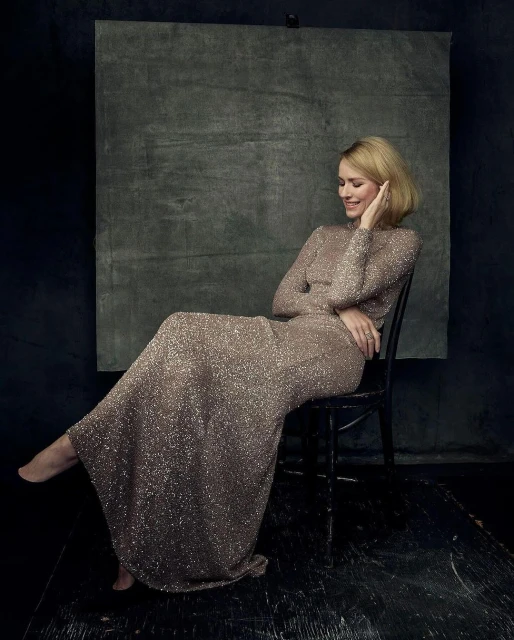 Adult female model in a shimmering long-sleeve gown seated on a black chair in a photo studio with a mottled grey backdrop.