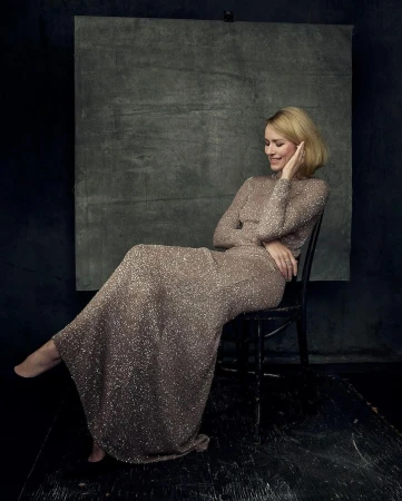 Naomi Watts in a long shimmering evening gown seated on a dark wooden chair in a studio.