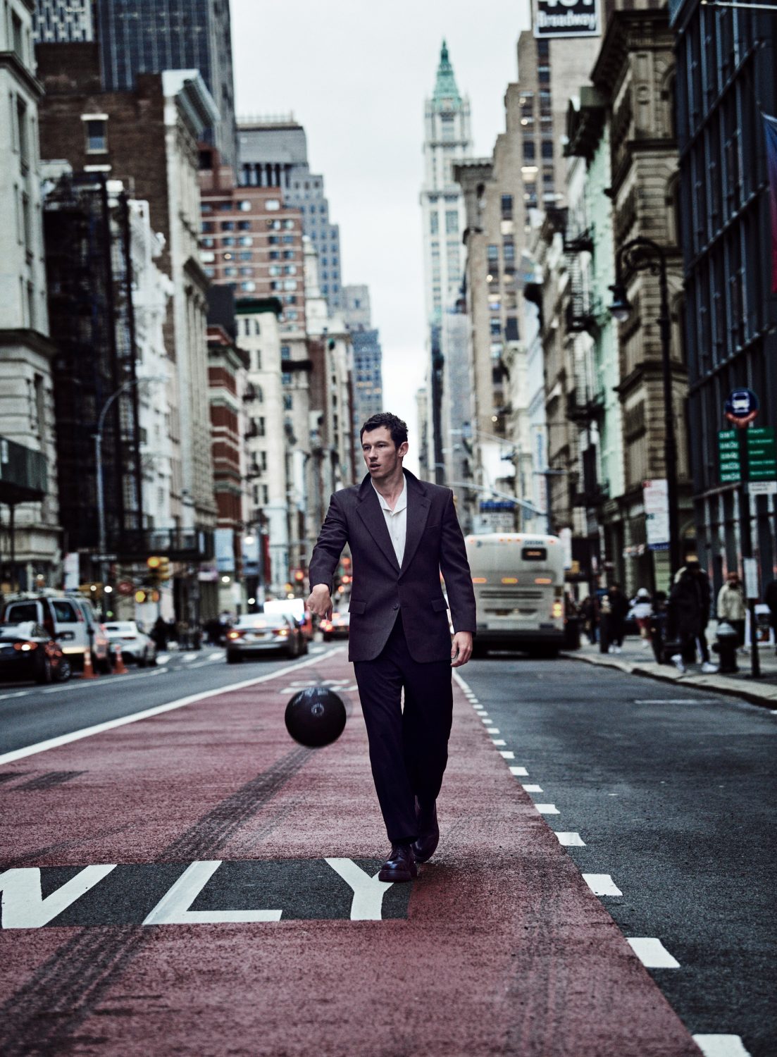 A man in a suit dribbles a basketball on an urban street with skyscrapers. He is walking toward the camera on a red painted crosswalk.