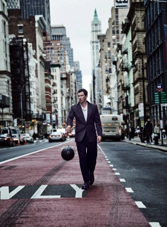 A man in a suit dribbles a basketball on an urban street with skyscrapers. He is walking toward the camera on a red painted crosswalk.