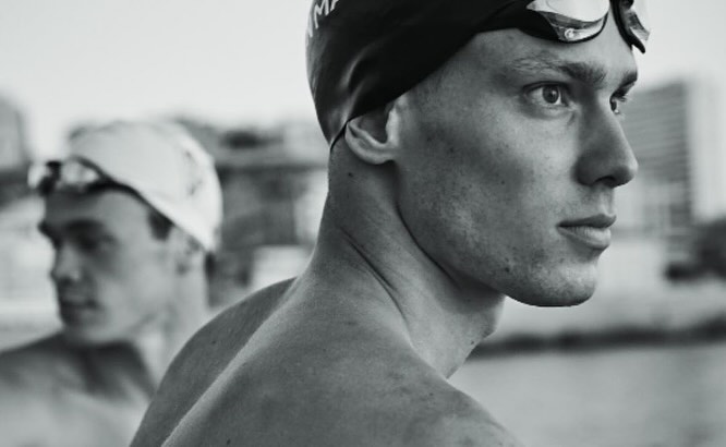 Monochrome close-up of two swimmers with caps and goggles, one in focus in the foreground and one blurred in the background.