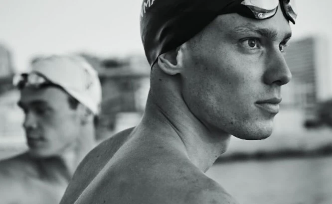 Monochrome close-up of two swimmers with caps and goggles, one in focus in the foreground and one blurred in the background.