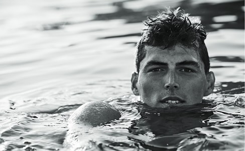 Black and white portrait of a young adult male swimmer in water, cropped at the shoulders, with a blurry water background.