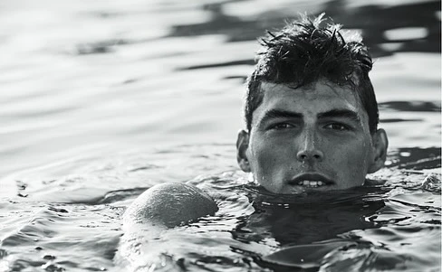 Black and white portrait of a young adult male swimmer in water, cropped at the shoulders, with a blurry water background.